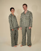 A man and a woman modeling matching striped pajama sets against a plain beige studio background. The pajamas feature vertical stripes in dark green and off-white, with a long-sleeve button-up top and matching pants.