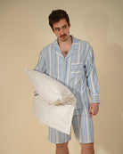 A man wearing a light blue and white vertically striped pajama set holds a white pillow against a solid beige studio background.