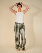 A model stands barefoot against a warm beige studio background, wearing a white sleeveless top and wide-leg trousers with vertical olive green and off-white stripes. The model has short dark hair and is holding it up with both hands.