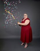 Woman in a red dress holding a bottle with confetti falling around her on a dark background. This dress is the Upton Dress & Skirt sewing pattern from Cashmerette.