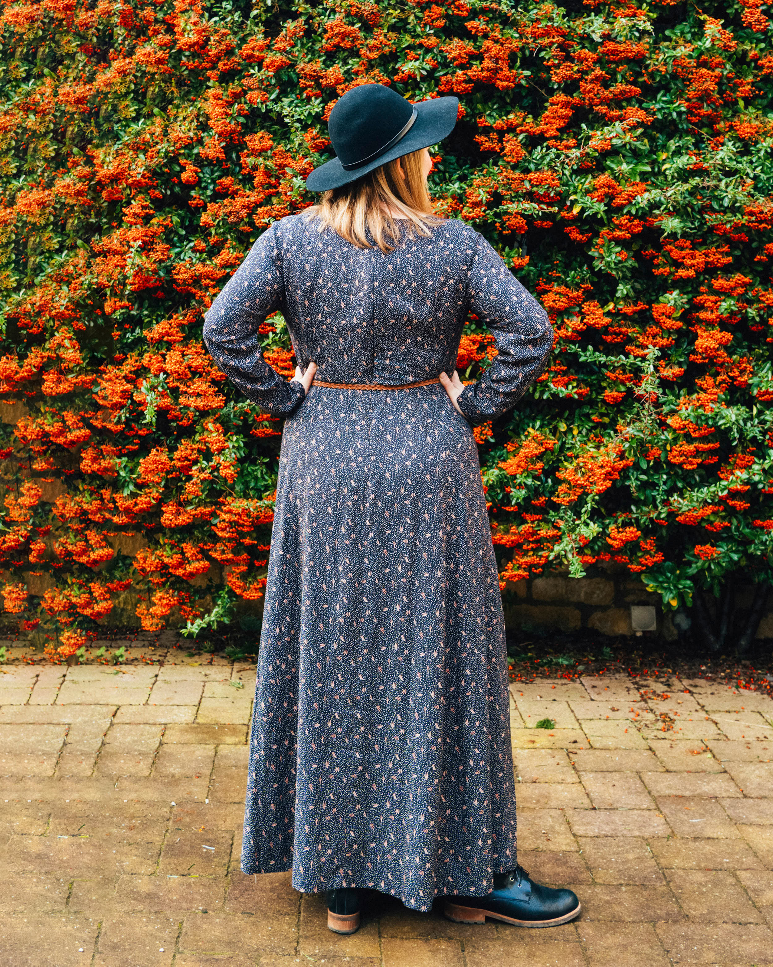 Woman in a long dark dress and black hat standing in front of a wall of orange flowers.. This dress is the Upton Dress & Skirt sewing pattern from Cashmerette.